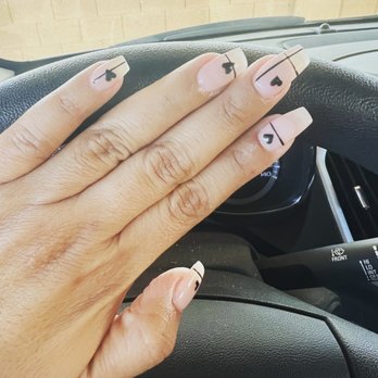 Uni Nails - A hand with neatly manicured nails is shown resting on a car steering wheel. The nails have a light pink base color with black designs, including hearts, arrows, and lines. The background includes part of the car's dashboard and windshield, indicating the photo was taken inside a vehicle. The nail art is simple and geometric, emphasizing minimalistic black motifs on a neutral background. - Nail Salons Near Me - Tempe, Arizona