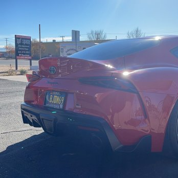 Body Master - Fixed - The image shows a red Toyota Supra parked outdoors on a paved surface. The car's rear end is visible, with a license plate reading BJDM649. The sky is clear and blue, with sunlight creating reflections on the car's surface. In the background, there are commercial signs and power lines, indicating a commercial or retail area. - Body Shops Near Me - Albuquerque, New Mexico