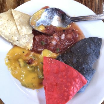 Frankie's at the Cassanova - free chips, salsa and queso - A white round plate contains tortilla chips, salsa, and multiple sauces, including a yellow cheese sauce and a red sauce. A metal spoon rests on the plate, partially covered with sauce. The plate is placed on a wooden surface, and the food appears to be Mexican cuisine. - New American Near Me - Pecos, New Mexico