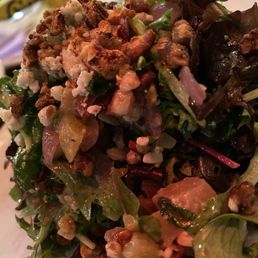 Beet Salad - A close-up image of a mixed green salad featuring different types of leafy greens, topped with chopped nuts and crumbled cheese. The salad appears fresh and is served on a white plate, with a blurred background suggesting a restaurant setting.