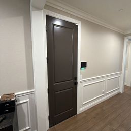 An interior hallway features a dark brown door with decorative panels, white wall paneling, and a digital keypad or control panel on the wall.