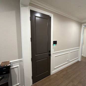 The Unhinged Door - An interior hallway features a dark brown door with decorative panels, white wall paneling, and a digital keypad or control panel on the wall. - Door Sales/Installation Near Me