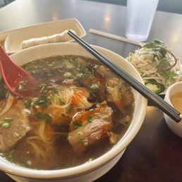 Spring roll, smoke brisket pho and a hot jasmine tea - A bowl of beef pho with herbs, noodles, and broth is on a table, accompanied by side dishes, chopsticks, and a cup of tea.