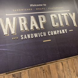 a sign for wrap city sandwich company - A wall sign features the name 'Wrap City' with details about sandwiches, wraps, and flatbreads, along with the establishment year 2015.