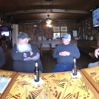 Docs Pub - The Burlington Motley Crew! - Four men are seated at a table in a pub with wooden walls and a brick accent wall, each with a beer bottle, in a casual setting. - Irish Pub Near Me - Burlington, New Jersey