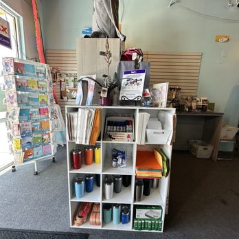 Beyond Mail and Print - The image shows the interior of a retail store with a white cubical shelving unit in the foreground, stocked with office supplies such as notebooks, folders, and thermoses in multiple colors. Behind the shelf, there is a counter with more products and a slatwall panel on the wall. To the left, a display rack holds greeting cards or similar items. The store has a carpeted floor, a window with a partial view outside, and a ceiling with visible wiring and lighting fixtures. - Printing Services Near Me - Rio Rancho, New Mexico