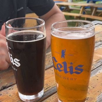Celis Brewery - Helles and dark - Two glasses of beer, one dark and one light, are positioned on a wooden table at Celis Brewery. The dark beer is in a tall glass with condensation, and the light beer is in a similar glass with the brewery's logo. A person wearing a black shirt is partially visible behind the glasses, and the background shows an outdoor seating area with picnic tables and a tent structure. - Breweries Near Me - Austin, Texas