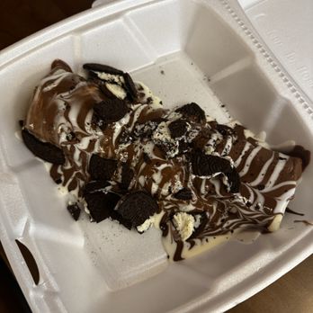 Crepe Stop - Crepe - A dessert crepe is presented in a foam takeout container, topped with crushed Oreo cookies, and drizzled with chocolate and white sauces. The crepe appears to be filled with a creamy substance, and the toppings are scattered unevenly across the surface. The container is placed on a wooden surface with some napkins visible in the background. - Creperies Near Me - Columbus, Ohio
