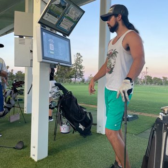 Dobson Ranch Golf Course - A man wearing a tank top, shorts, and a cap stands at a golf driving range, observing a screen with golf information. - Golf Near Me - Mesa, Arizona