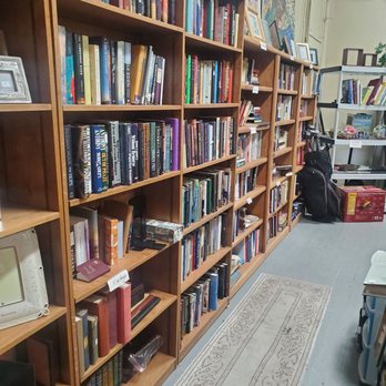 Serendipity Shoppe - Multiple wooden bookshelves are filled with books of various sizes and colors, arranged vertically. The shelves are positioned along a wall in an indoor space, with framed pictures and artwork displayed on top. A narrow rug runs along the floor in front of the shelves, and additional shelving units and items are visible in the background, indicating a retail or donation setting. - Thrift Stores Near Me - Buffalo, New York