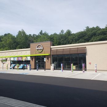 Nouria - The image shows the exterior of a Nouria convenience store located in Windham, NH. The store has a contemporary design with large glass windows, a prominent Nouria sign above the entrance, and a small brown accent wall. In front of the store, there are several parking spaces, including a designated handicapped spot, with a paved lot extending to the street. The background includes a wooded area with lush green trees under a partly cloudy sky. - Convenience Stores Near Me - Windham, New Hampshire