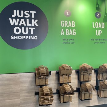 Amazon Fresh - Brown paper shopping bags are organized in racks on a light-colored floor, with a green wall featuring signage about shopping procedures. - Grocery Near Me - Oceanside, New York