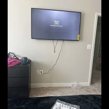 FixTman - Washington - A wall-mounted flat-screen television displays a 'Downloading' message on the screen. The TV is connected to a power outlet via a visible cord hanging down. To the left, there is a black dresser with some items on top, including a pink bag and a black object. The room has beige carpet flooring, and a doorway is visible on the right side of the image. A piece of paper or packaging is partially visible in the foreground. - Furniture Assembly Near Me