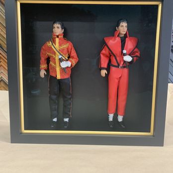 The Great Frame Up - The shadow box contains two Michael Jackson dolls dressed in iconic red and black outfits. The doll on the left wears a red jacket with gold embroidery, black pants, and white gloves, holding a microphone. The doll on the right is dressed in a red jumpsuit with black accents, also holding a microphone. The display is set against a black background with a gold trim around the frame, and the background features a pattern of various picture frame samples. - Framing Near Me - Spring, Texas