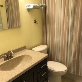 Home Fixers - Before photo - The image shows a bathroom with a dark wood vanity cabinet topped with a beige countertop and an integrated sink. A mirror is mounted on the wall above the vanity. Next to the vanity, there is a toilet positioned near a striped fabric shower curtain. A towel is folded on a shelf above the toilet, and the bathroom has light-colored walls and tiled flooring. - Handyman Near Me - Winfield, Illinois