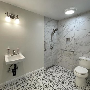 Westco Development - Bathroom conversion to ADA compliant roll in shower. - Bathroom features a walk-in shower with marble tiles, a small wall-mounted sink, and patterned floor tiles, with lighting fixtures on the ceiling. - General Contractors Near Me - Glendora, California