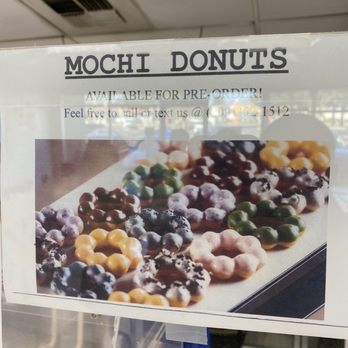 Sunshine Donuts - a sign for a donut shop - The image shows a sign for a donut shop named 'Mochi Donuts,' featuring a photograph of colorful mochi donuts with different toppings. The sign indicates that the donuts are available for pre-order and provides a phone number for calls or texts. The background includes a ceiling with a vent, suggesting the sign is placed inside or near the shop. - Donuts Near Me - Turlock, California