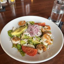 Grilled Shrimp Salad - A shrimp salad featuring grilled shrimp, avocado slices, tomato wedges, and sliced red onion on a white plate, with a glass of water nearby.