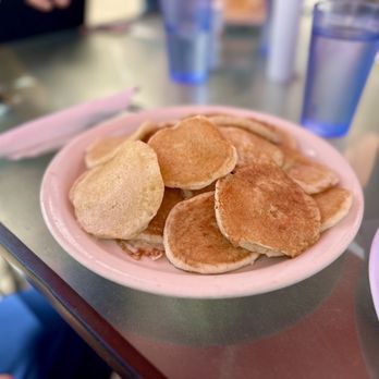 Joes Cafe - Pancakes- Taste great! - A pink ceramic plate contains several small pancakes with a golden-brown color, arranged on a table. The table has various items including blue-tinted glasses, utensils, and napkins, indicating a dining setting. The background is slightly blurred, showing other table elements and possibly other diners. - Breakfast & Brunch Near Me - Orem, Utah