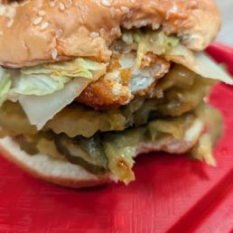 Buffalo Chicken Sandwich - A close-up image of a partially eaten chicken sandwich featuring a sesame seed bun, lettuce, pickles, and sauce. The sandwich appears to contain fried chicken, with visible layers of vegetables and condiments. It is served on a red tray, with a blurred background indicating an indoor dining setting.