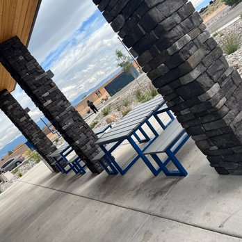 Dutch Bros Coffee - An outdoor seating area features stone pillars supporting a wooden roof, with picnic tables and benches on a concrete surface, and a parking lot in the background. - Coffee & Tea Near Me - Albuquerque, New Mexico