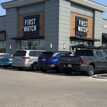 First Watch - Exterior - The photo depicts the front exterior of a First Watch restaurant, featuring a modern building design with gray and wood accents. The signage displays the First Watch logo prominently. Multiple vehicles, including a white minivan, a blue SUV, and a gray pickup truck, are parked in the parking lot in front of the restaurant. The sky is clear and blue, indicating a sunny day. - Breakfast & Brunch Near Me - Naperville, Illinois