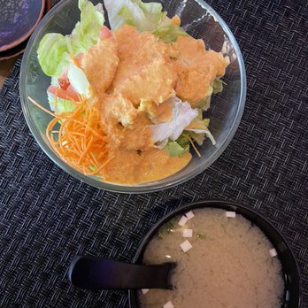 Akari Sushi - a bowl of rice and a bowl of soup - The image shows a clear glass bowl containing a salad with lettuce, shredded carrots, and a generous amount of salad dressing on top. Next to the salad, there is a black bowl of miso soup with small cubes of tofu and green onions floating on the surface. The items are placed on a black textured placemat, with part of a dark wooden table visible. - Sushi Bars Near Me - Poughkeepsie, New York