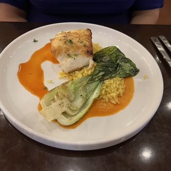 Red Sage - Chilean Sea Bass - The image shows a white round plate with a serving of cooked fish, orzo pasta, and vegetables, including a piece of leafy green and a piece of fennel. The fish is topped with herbs and placed on a bed of orzo, with a reddish-orange sauce surrounding the food. The plate is on a dark wooden table, with utensils and a smartphone visible nearby. - New American Near Me - Santa Fe, New Mexico