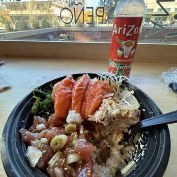 Aloha Cones - 2 combo with sesame ahi and salmon ( sorry I had already taken a few bites before deciding I need to document it) - A poke bowl containing slices of salmon, tuna, rice, and vegetables, with a bottled Arizona watermelon-flavored water, placed on a wooden table by a window. - Poke Near Me - Denver, Colorado