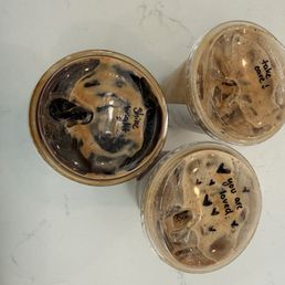 Toffee - Three iced coffee drinks with foam and ice are arranged on a white surface. Each cup has handwritten messages on the lids, including 'Shine bright,' 'take love,' and 'you are loved,' with small heart symbols. The drinks appear to be from a café, featuring dark coffee with foam and ice cubes visible through the clear plastic cups.