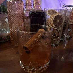 old fashioned - A glass of old fashioned cocktail with a cinnamon stick inside is placed on a wooden bar counter. Behind it, there are bottles of bitters and other ingredients, along with a container holding dried lemon slices. The background shows a person and a dimly lit bar setting, indicating a social drinking environment.