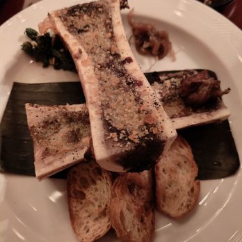 CODE RED - Bone marrow - A dish featuring bone marrow served on a white plate, garnished with toasted bread, greens, and condiments. The marrow is topped with seasoning or breadcrumbs, and a small portion of a side or garnish is visible. A dark sauce is in a bowl nearby, and the presentation suggests a fine dining or specialty restaurant setting. - Cocktail Bars Near Me - Washington, DC, District of Columbia