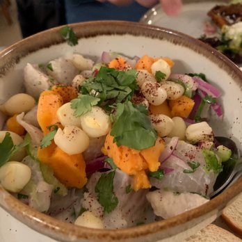 Roots Latin Grill - Peruvian ceviche - A ceramic bowl contains Peruvian ceviche with pieces of fish, corn, sweet potato, red onion, and cilantro. The dish is garnished with herbs and spices, and a spoon is partially visible. In the background, there is another plate with food, possibly a different dish, on a table setting. - Latin American Near Me - Springfield, Illinois