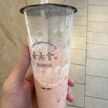 TP Tea - strawberry green tea - A person holds a transparent plastic cup containing pink strawberry milk tea with foam on top. The cup has black plastic sealing film and printed text, including Chinese characters and the words 'Taiwan Professional Tea' and 'Since 2005.' The background shows a cafe interior with tiled flooring, a wooden wall, and a person wearing sandals and light-colored pants sitting at a table. - Bubble Tea Near Me - Flushing, New York