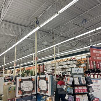 Hobby Lobby - The image shows the interior of a retail store, likely a craft or general merchandise store, with high ceilings and bright fluorescent lighting. Shelves are filled with holiday decorations, including plush snowmen, Christmas-themed items, and greeting cards. A customer is seen browsing the card display, and promotional signs are visible, indicating sales and exchange policies. The store appears organized and well-lit, with various seasonal products on display. - Home Decor Near Me - Layton, Utah