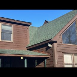 New roof - The image shows a two-story house with brown wooden siding and a green shingled roof. The house has multiple windows, including a large triangular window on the upper floor. A small porch area is visible at the front, with a light fixture and a wind chime hanging. The sky above is clear and blue, and part of a neighboring roof can be seen in the background.