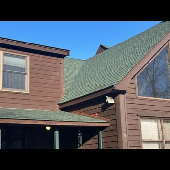 Gannon Construction - New roof - The image shows a two-story house with brown wooden siding and a green shingled roof. The house has multiple windows, including a large triangular window on the upper floor. A small porch area is visible at the front, with a light fixture and a wind chime hanging. The sky above is clear and blue, and part of a neighboring roof can be seen in the background. - General Contractors Near Me - Scituate, Massachusetts
