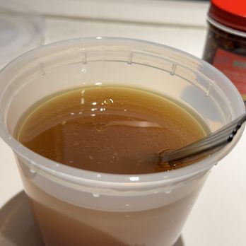 Roll Play - Side Pho - A clear plastic cup filled with tea, with a metal spoon inside, on a white table. A jar with a red lid is visible in the background. - Vietnamese Near Me - Vienna, Virginia