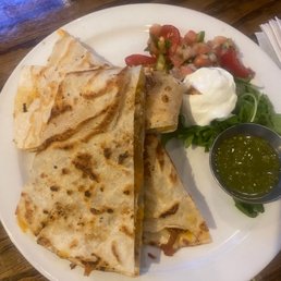 Chicken Tinga Quesadilla - A plate features a quesadilla cut into four triangular pieces, with a golden-brown, toasted exterior. It is served with a side of salsa, a dollop of sour cream, and some fresh greens. The dish is presented on a white plate placed on a wooden table, with a small container of green salsa or sauce on the side.