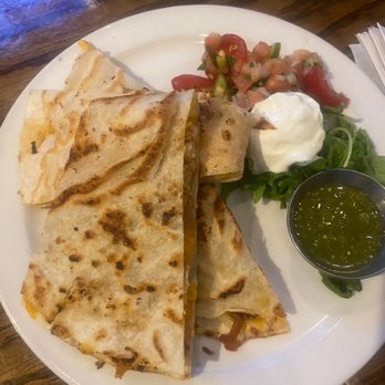 The Globe Pub - Chicken Tinga Quesadilla - A plate features a quesadilla cut into four triangular pieces, with a golden-brown, toasted exterior. It is served with a side of salsa, a dollop of sour cream, and some fresh greens. The dish is presented on a white plate placed on a wooden table, with a small container of green salsa or sauce on the side. - Sports Bars Near Me - Chicago, Illinois