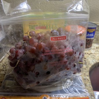 ALDI - $2 - A transparent plastic bag filled with red seedless grapes is positioned on a kitchen countertop. The bag has a red label with the text 'PLU #4023' and indicates the grapes are produce of Mexico. In the background, there are canned beans and a power outlet on a beige wall. The surface of the countertop is speckled with various shades of brown and beige. - Grocery Near Me - Slidell, Louisiana