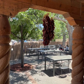 Village Pizza - A bunch of red chili peppers is suspended from a wooden frame in an outdoor dining area. The setting includes several tables and chairs, shaded by trees with green leaves. The background shows a wooden fence, parked cars, and a few people sitting at tables. The scene is illuminated by sunlight, creating shadows on the ground. - Pizza Near Me - Corrales, New Mexico