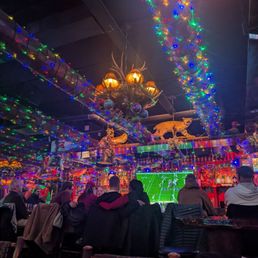 The image shows a group of people seated at a bar, watching a football game on a large screen. The bar is decorated with multicolored Christmas lights and holiday-themed ornaments, including a chandelier with antlers and ornaments hanging from the ceiling. The setting appears festive, with various decorations and a cozy atmosphere suitable for social gatherings during the holiday season.
