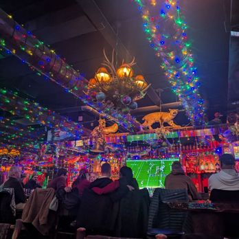 Camp Bar Third Ward - The image shows a group of people seated at a bar, watching a football game on a large screen. The bar is decorated with multicolored Christmas lights and holiday-themed ornaments, including a chandelier with antlers and ornaments hanging from the ceiling. The setting appears festive, with various decorations and a cozy atmosphere suitable for social gatherings during the holiday season. - Bars Near Me - Milwaukee, Wisconsin
