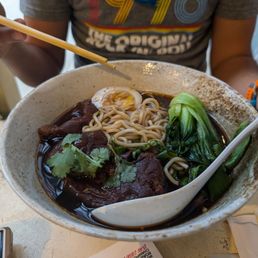 Braised Beef Noodles Soup - A bowl of ramen featuring braised beef, leafy greens, a soft-boiled egg, and noodles in a dark broth, with chopsticks and a spoon.