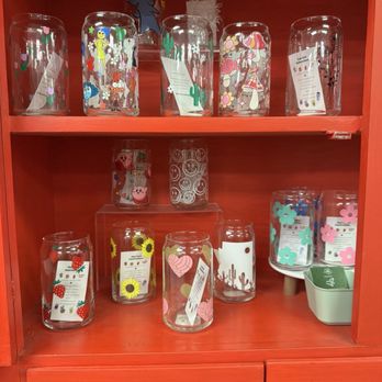 Junk Head - Cute glass tumblers! - Multiple glass tumblers featuring colorful designs such as characters, flowers, and abstract patterns are displayed on a red wooden shelf. Some tumblers are placed directly on the shelf, while others are elevated on small stands. A small container with additional tumblers is positioned on the right side of the shelf. The background includes a decorative element with a sun illustration and a cartoon character, contributing to a lively and decorative display. - Used, Vintage & Consignment Near Me - Mesa, Arizona
