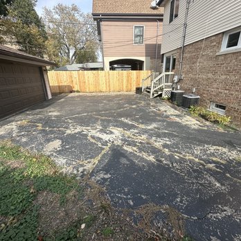 Prestigious Pavement - Before - driveway in terrible shape - A large asphalt driveway with extensive cracking and peeling, bordered by a wooden fence and adjacent to residential buildings. - Masonry/Concrete Near Me - Alsip, Illinois