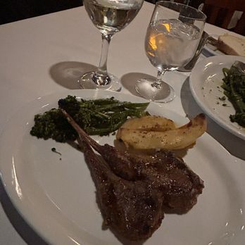 Avli Estiatorio - Lamb chops(I split with another guest in our party) - A dinner plate features lamb chops, sautéed broccoli rabe, and potato wedges, with two glasses of water and a glass of white wine on a white tablecloth. - Greek Near Me - Winnetka, Illinois