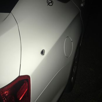 Kitu's Auto - Bullet hole - The image depicts a white car parked outdoors at night, with a visible bullet hole in the rear quarter panel. A key is placed on the roof, and a charging cable is connected to the vehicle. The photo is taken in low light, highlighting the damage and objects on the car's surface. - Body Shops Near Me - Cincinnati, Ohio