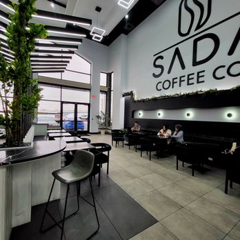 Sada Coffee - The image shows the interior of Sada Coffee Co., featuring high ceilings with modern geometric lighting fixtures. The space includes a seating area with black chairs and tables, and a large logo on the wall reading 'Sada Coffee Co.' The front entrance has glass doors and windows, allowing natural light to illuminate the space. There are a few customers seated, and the decor includes plants and minimalist design elements. - Desserts Near Me - Palos Hills, Illinois